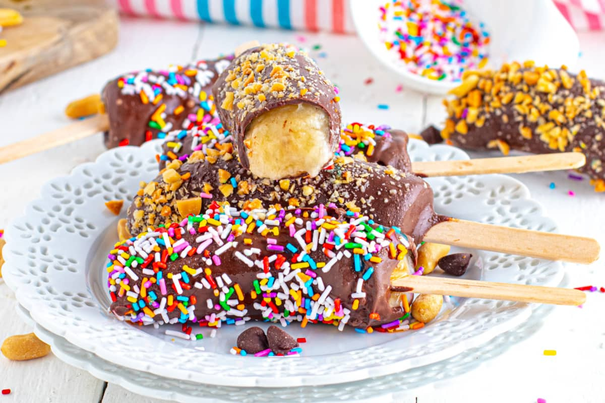 Handheld chocolate-covered frozen bananas with sprinkles and nuts on a white plate.