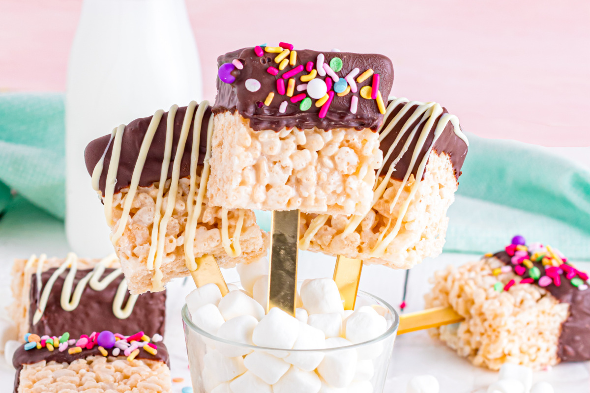 Rice krispie treats on a stick, dipped in chocolate and sprinkled with colorful toppings, are handheld desserts displayed in a cup with marshmallows.