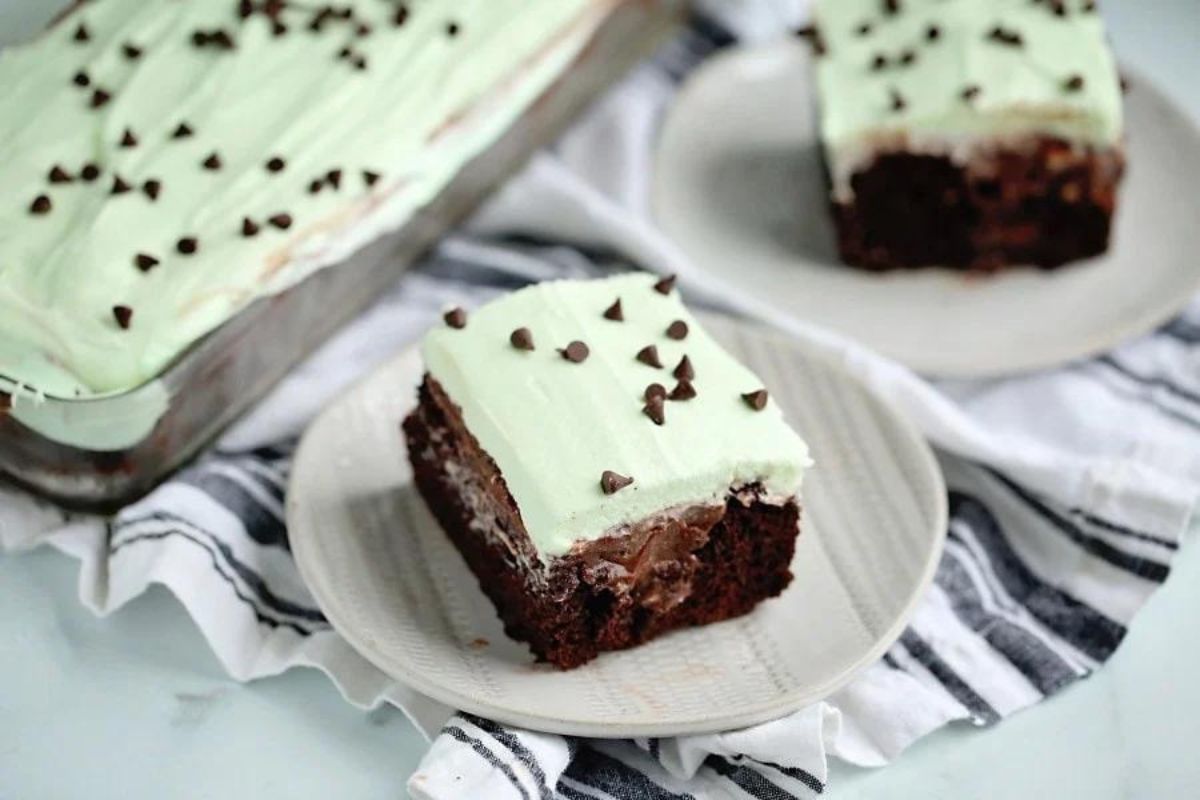 A chocolate poke cake with mint frosting and chocolate chip garnish, served on a plate with more cupcakes in the background.