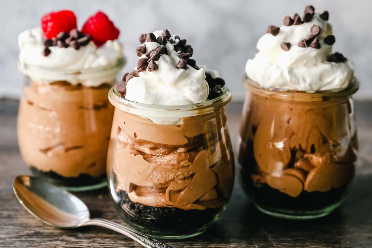 Three jars of chocolate mousse topped with whipped cream, chocolate chips, and raspberries, beside a spoon—perfect single-serve individual desserts for any occasion.