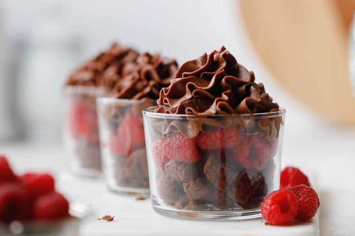 Three glass cups filled with raspberries, chocolate cake pieces, and chocolate whipped cream offer delightful single-serve individual desserts, with extra raspberries nearby.