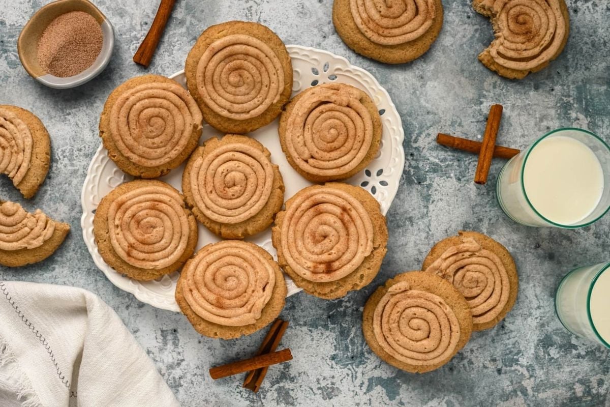 A plate of cinnamon swirl cookie recipes accompanied by a glass of milk, with extra cookies scattered around and cinnamon sticks for decoration.
