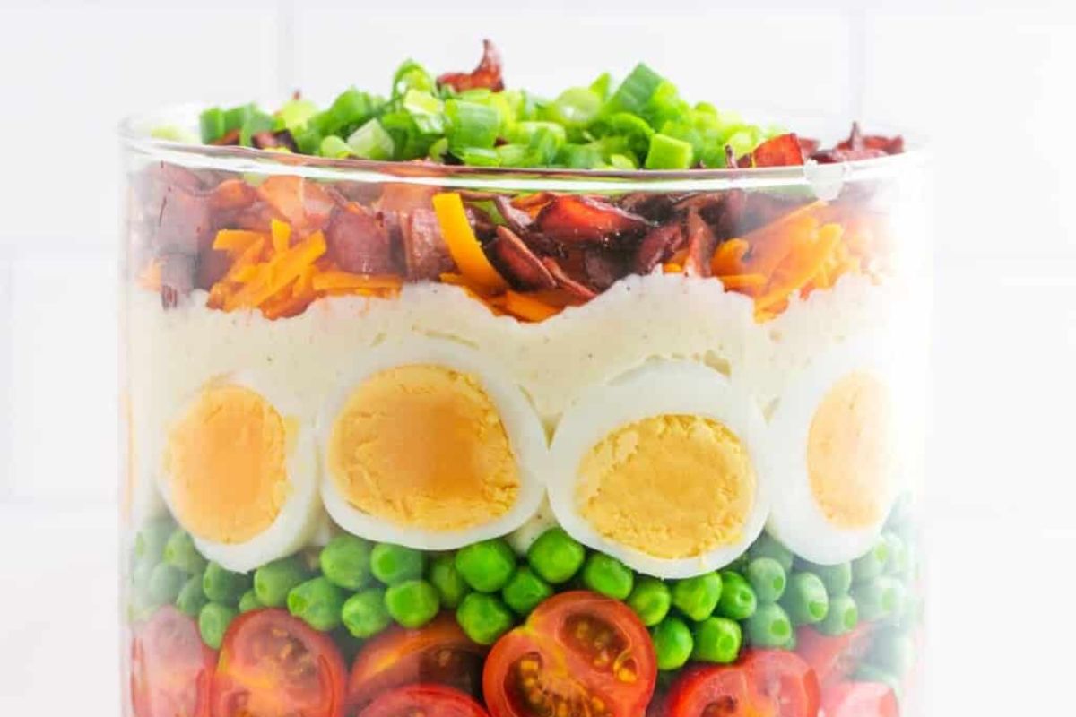 Layered salad with eggs, peas, tomatoes, bacon, and cheese in a clear bowl, perfect for Easter sides.