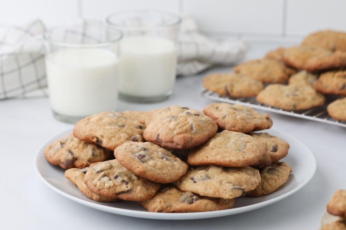 A plate of chocolate chip cookies, prepared from our finest cookie recipes, with additional cookies and two glasses of milk in the background.