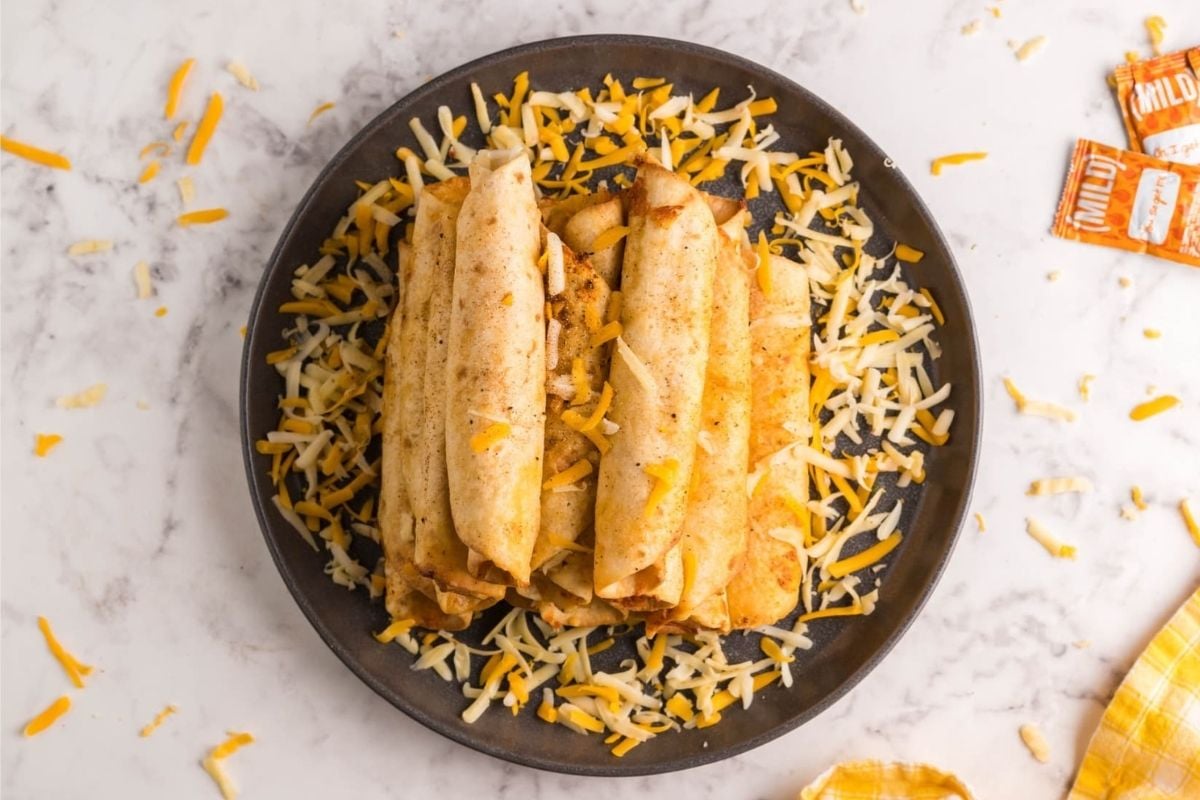 Three rolled tacos on a plate sprinkled with shredded cheese, perfect for cheesy recipes.