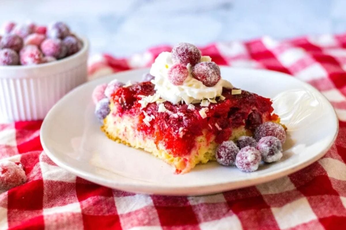 A slice of cranberry upside down cake, topped with whipped cream and sugared cranberries, sits on a white plate.