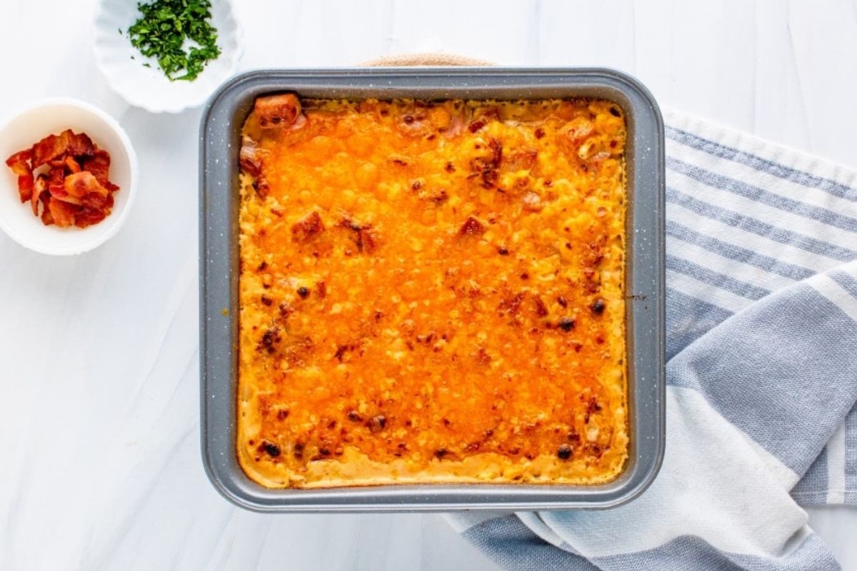 A freshly baked macaroni and cheese in a square baking dish on a wooden surface with a side of chopped herbs and bacon bits, perfect for those who love cheesy recipes.
