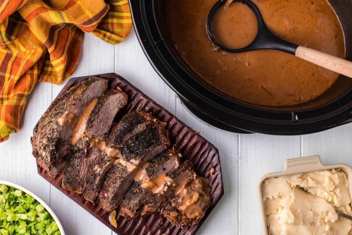 Sliced roast beef with gravy, mashed potatoes, and chopped greens—easy comfort food inspired by classic slow cooker recipes—served on a light table with a plaid napkin.