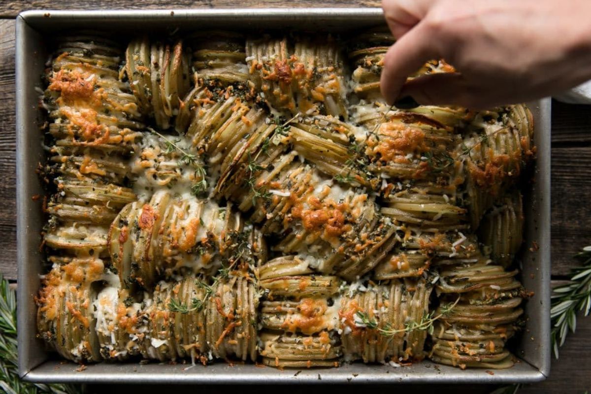A tray of baked hasselback potatoes, perfect as Easter sides, garnished with herbs, with a hand adding final touches.