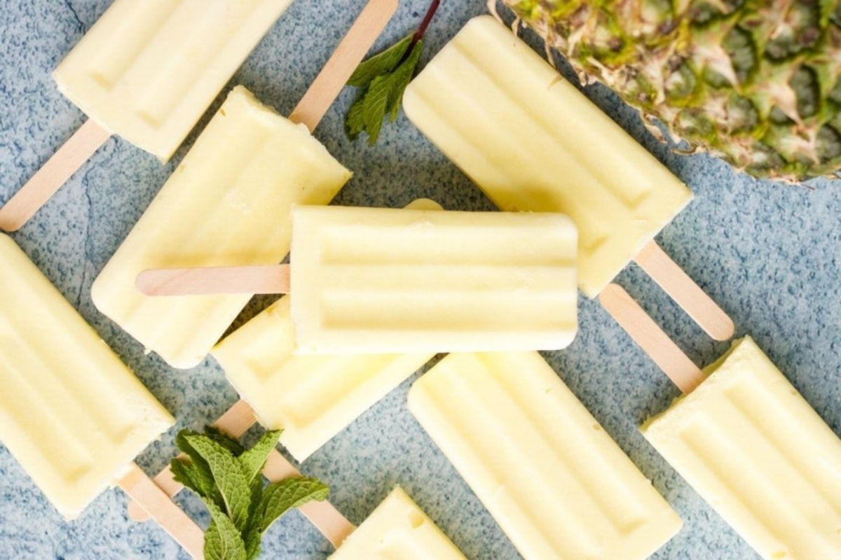 Homemade pineapple popsicle recipes on a blue surface with mint leaves and pineapple in the background.