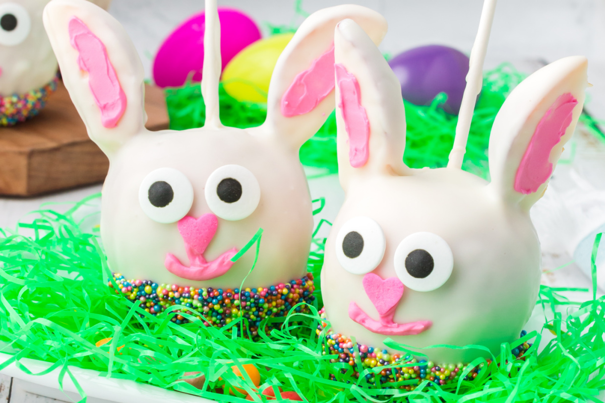 Two easter-themed Oreo dessert cake pops decorated to resemble bunnies, placed on a bed of colorful artificial grass with easter eggs in the background.