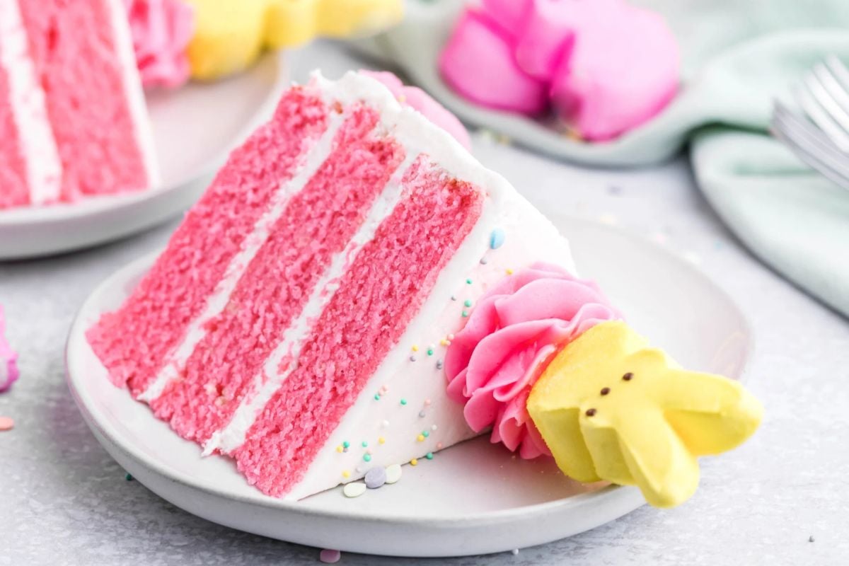 A slice of pink Easter cake with white frosting and colorful sprinkles, garnished with a piped flower and leaf on a white plate.