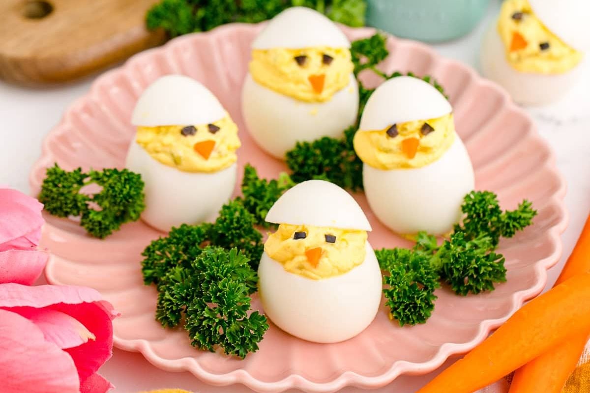 Deviled eggs creatively decorated to look like chicks, surrounded by parsley and carrot garnishes on a pink plate, serving as delightful Easter sides.