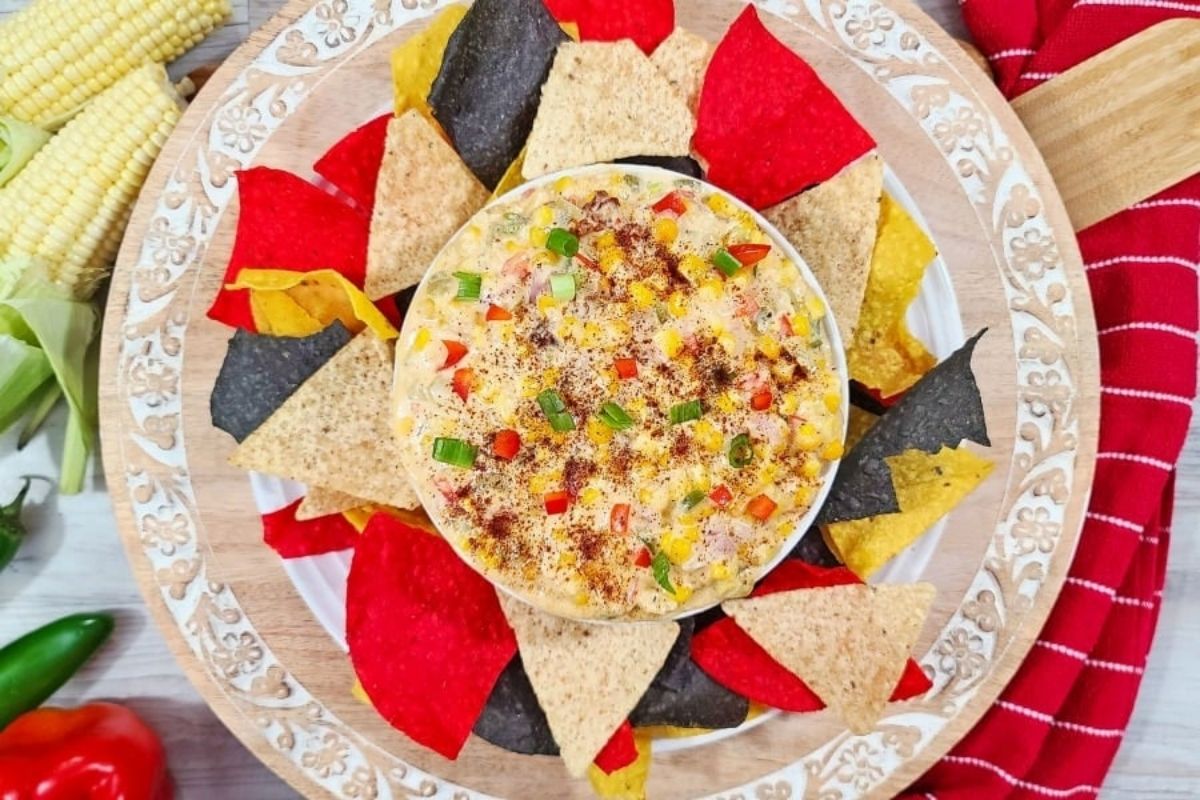 A bowl of colorful cheesy corn dip surrounded by a variety of tortilla chips on a wooden platter.