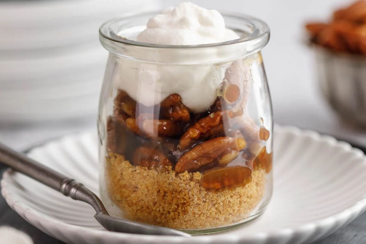 A single-serve glass jar with a crumb crust, pecans in syrup, and whipped cream on top, served on a white plate with a spoon—perfect for those who love individual desserts.