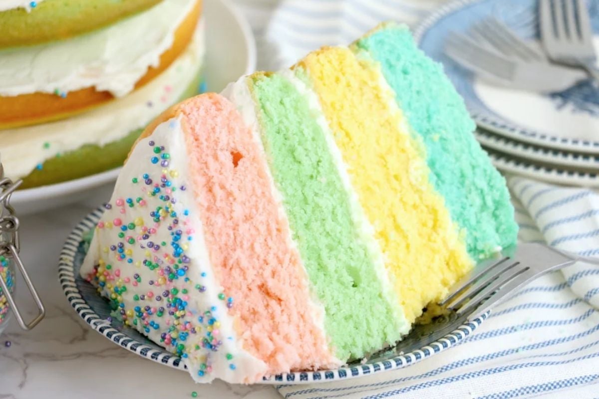 A slice of colorful Easter cakes with pastel pink, orange, green, and yellow layers, topped with white icing and sprinkles, served on a decorative plate.