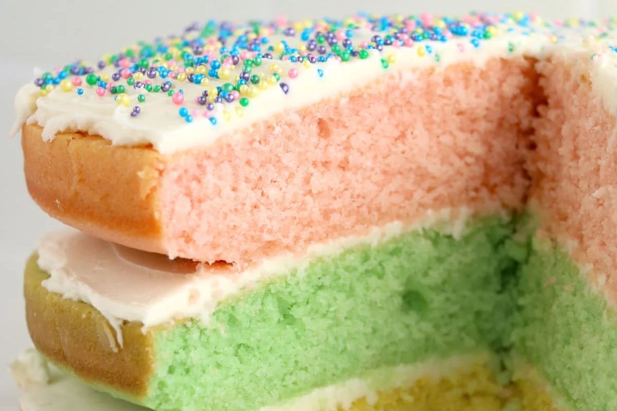 A close-up of a layered Easter cake with pink and green sponges, white frosting, and colorful sprinkles on top.