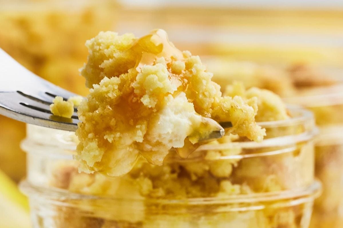 A fork holds a bite of crumbly cheesecake topped with honey, over a glass jar of single-serve individual dessert.