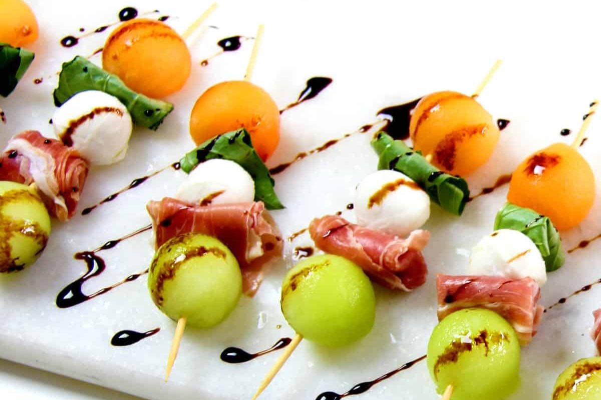 An arrangement of melon and prosciutto appetizers with balsamic glaze on a white plate, perfect for an Easter appetizer.