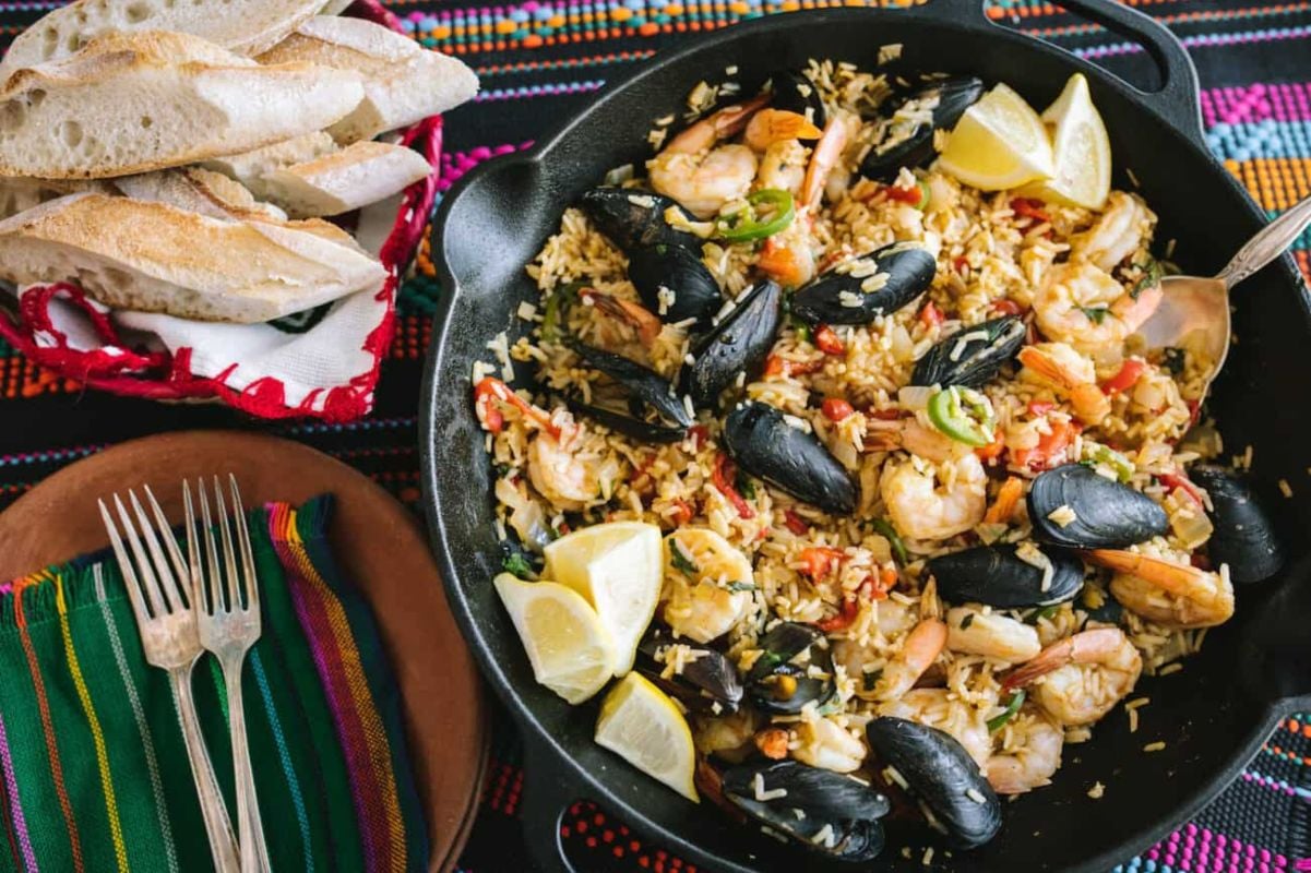 A skillet of seafood paella with shrimp, mussels, and lemon wedges, inspired by traditional seafood recipes, served next to a basket of bread and a fork on a colorful woven tablecloth.