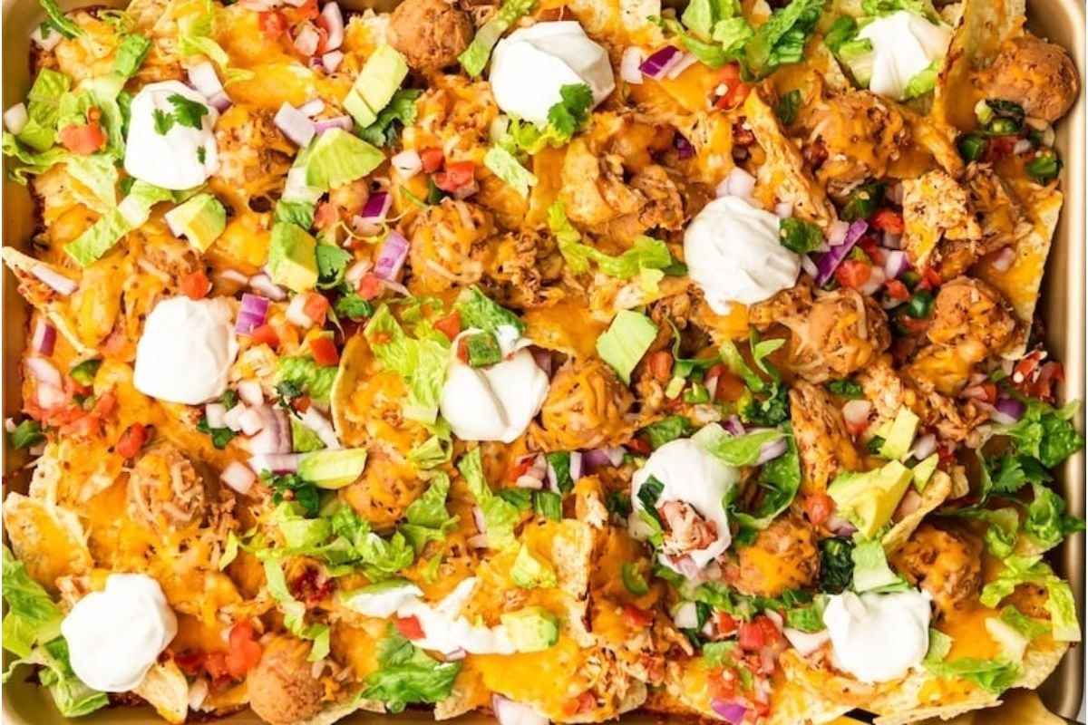 A tray of cheesy loaded nachos topped with shredded cheese, chicken, diced tomatoes, red onions, lettuce, and dollops of sour cream.