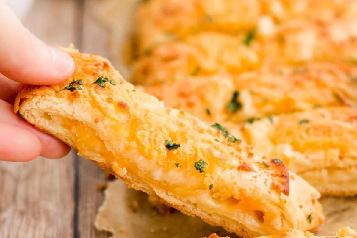 A hand holding a cheesy breadstick topped with herbs, with more breadsticks in the background—a perfect treat for fans of cheesy recipes.