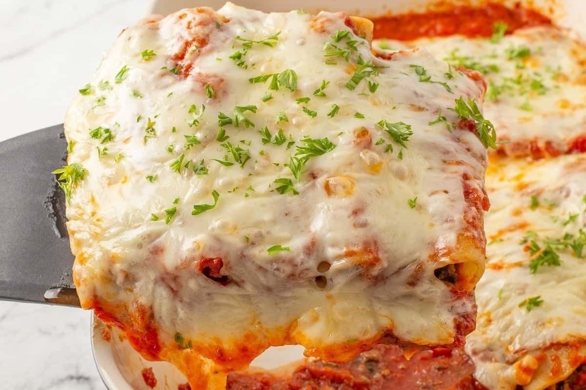 A lasagna slice being lifted from a baking dish, showing layers of pasta, meat, cheese, and tomato sauce, topped with melted cheese and parsley in this cheesy recipe.