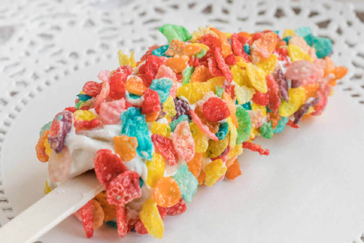 Handheld colorful cereal-coated popsicle on a white lace doily.