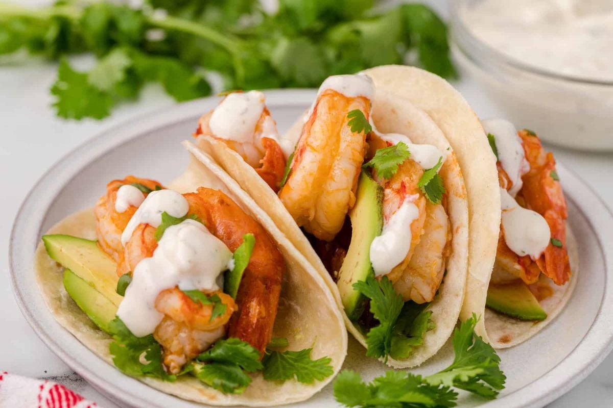 Three seafood recipes featuring shrimp tacos with avocado slices and cilantro in soft shells, garnished with cream sauce, served on a white plate.
