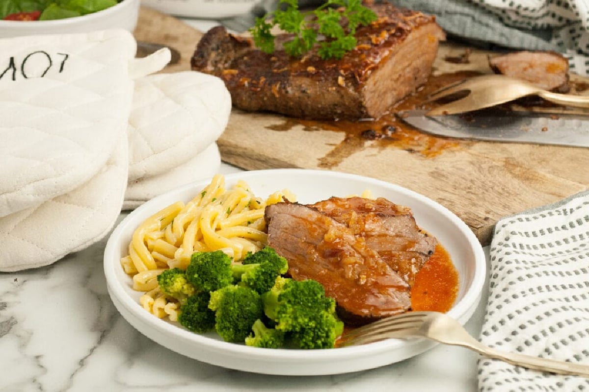 A plate of food with sliced beef, broccoli, and noodles from grandma's recipes, alongside a cutting board with a larger piece of cooked beef and utensils.