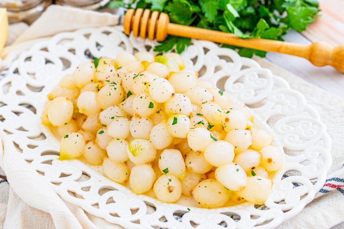 A plate of seasoned pearl onions garnished with herbs, accompanied by a honey dipper, inspired by recipes from grandma.