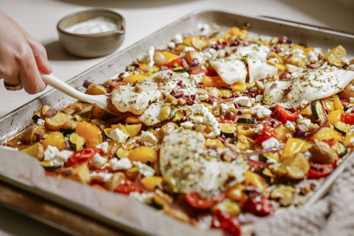 A baking sheet filled with roasted vegetables and white fish fillets, garnished with herbs, on a kitchen counter, perfect for seafood recipes.