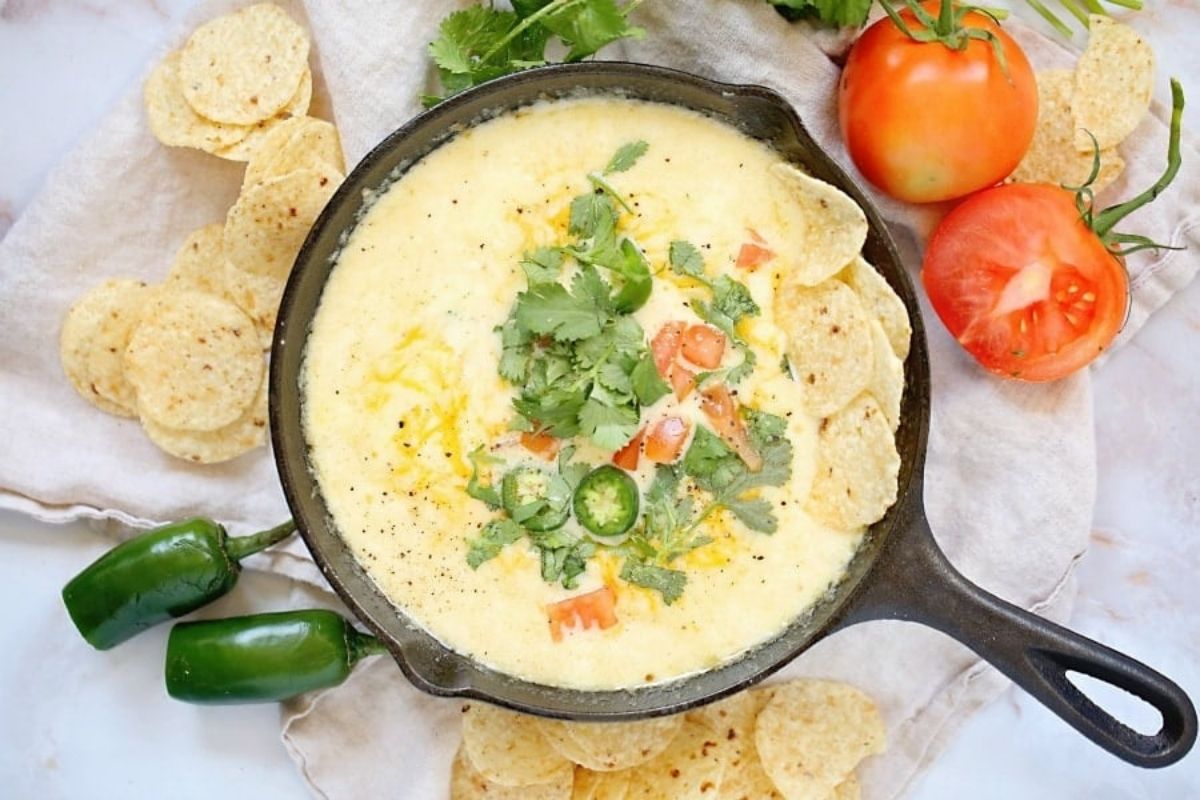 Cast iron skillet with warm, cheesy queso dip garnished with cilantro and jalape&ntilde;o, surrounded by tortilla chips and fresh tomatoes.