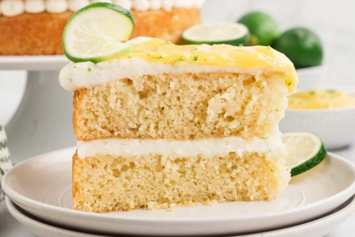 A slice of lime-flavored layer cake with white frosting on a plate, garnished with a lime slice and zest.