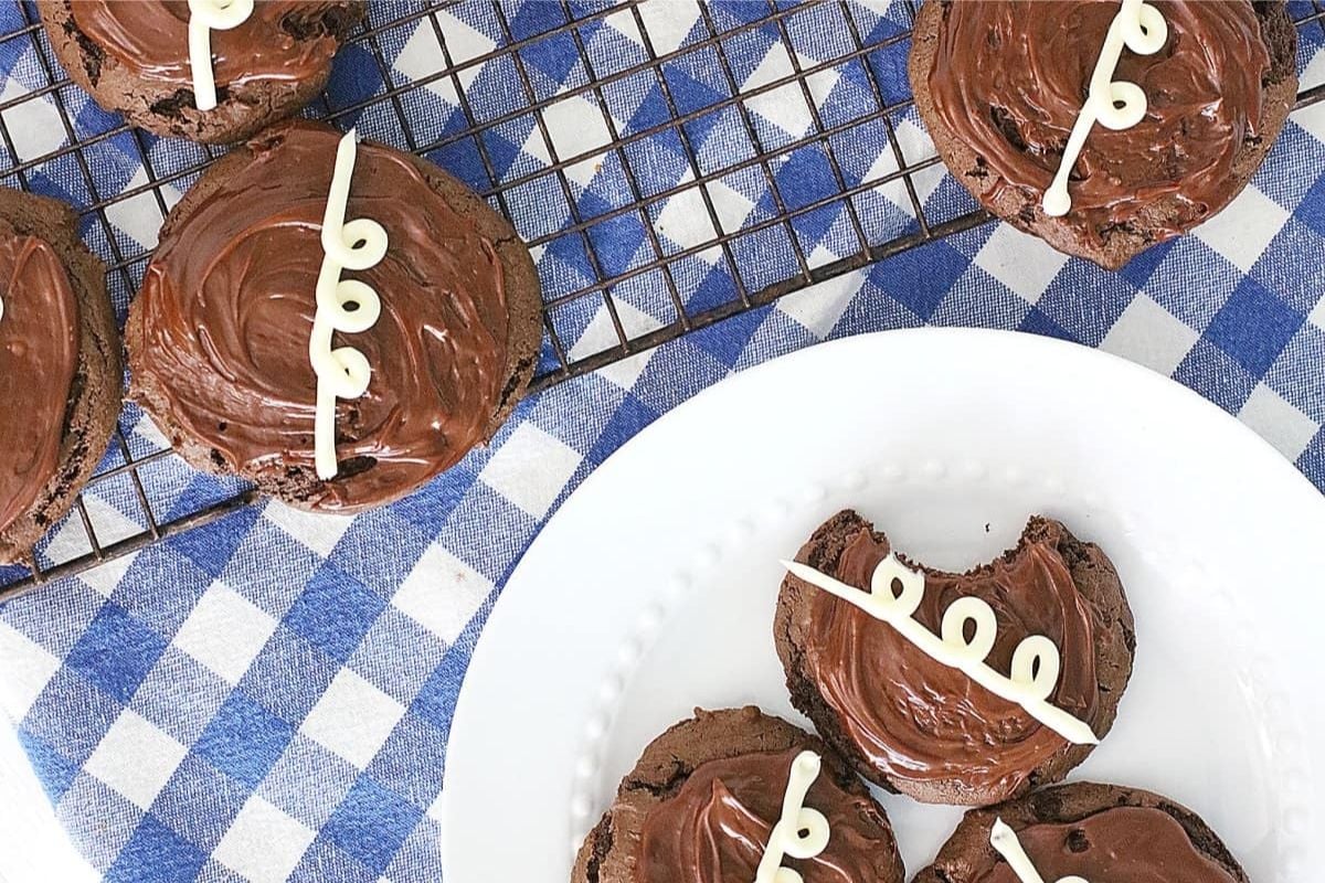 Chocolate frosted cookie recipes on a wire rack with a few on a plate, decorated with white icing designed to resemble football laces.