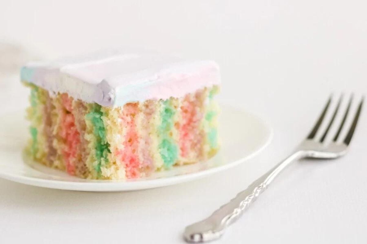 A slice of pastel rainbow poke cake with white icing on a plate beside a fork.