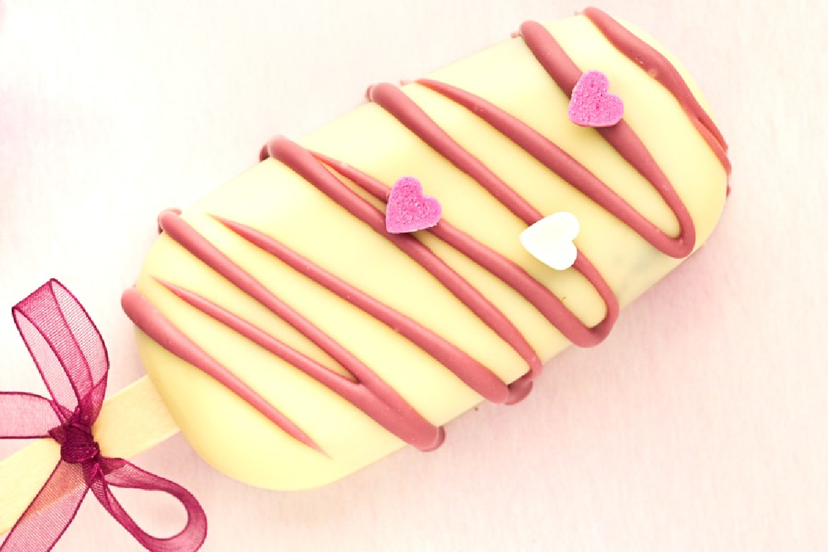 A decorated handheld eclair with pink icing and heart-shaped sprinkles on a pastel background.