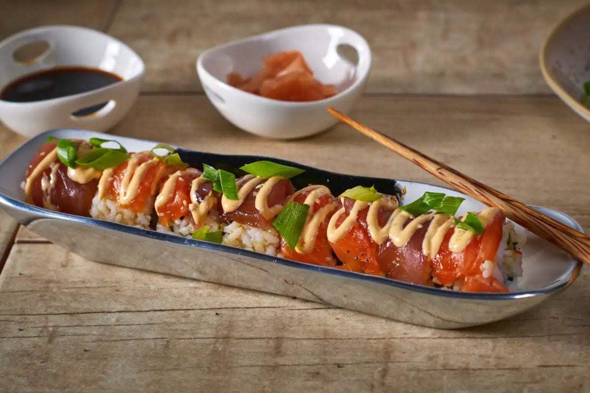 A plate of sushi rolls topped with avocado and drizzled with sauce, accompanied by soy sauce and pickled ginger on a wooden table, showcasing delightful seafood recipes.