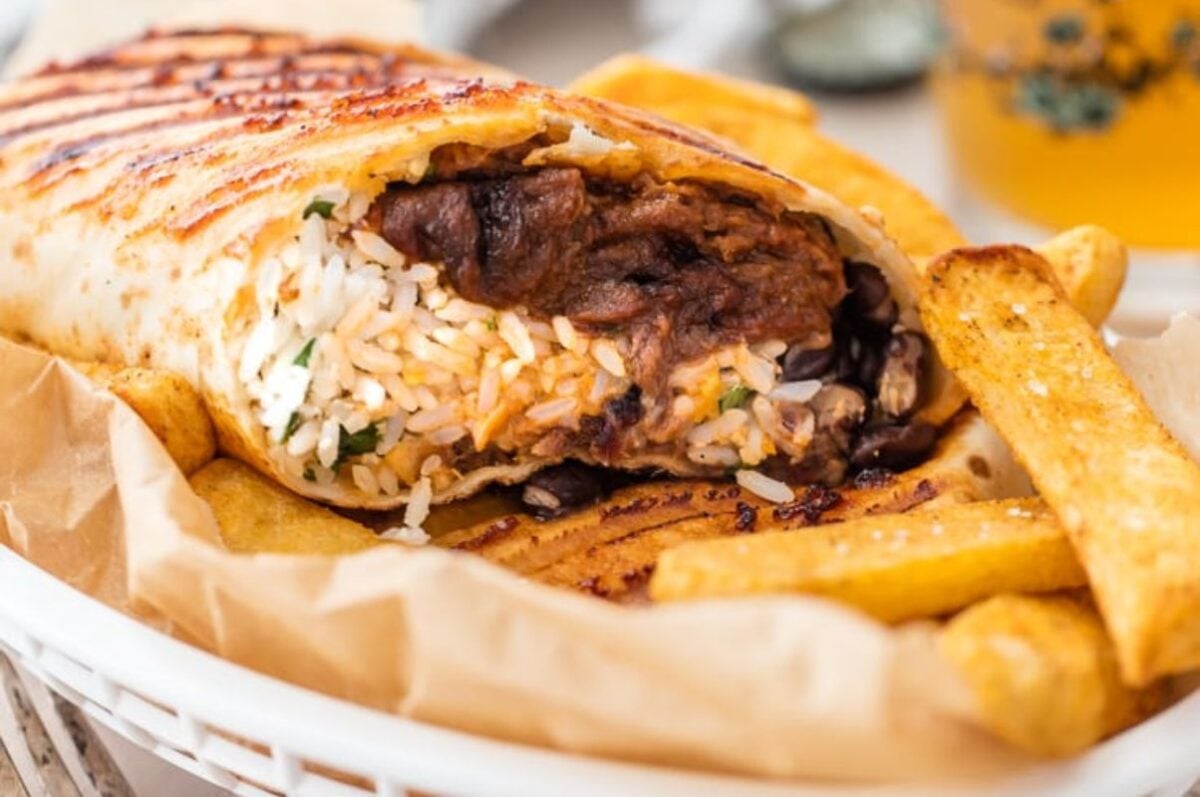 Close-up of a grilled burrito recipe filled with rice, beans, and meat, served with a side of golden-brown french fries on a wooden plate.