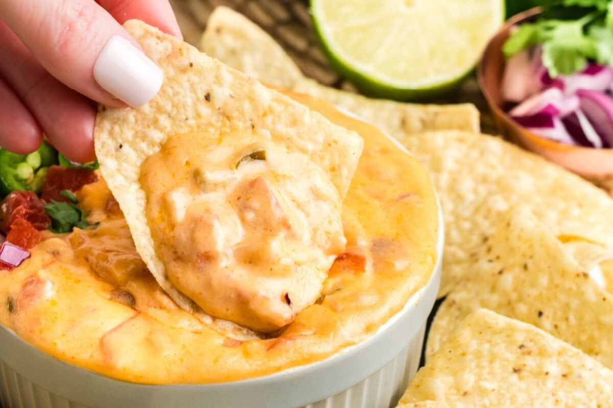 A hand dipping a tortilla chip into a bowl of creamy cheese dip with lime and cilantro in the background, perfect for an Easter appetizer.