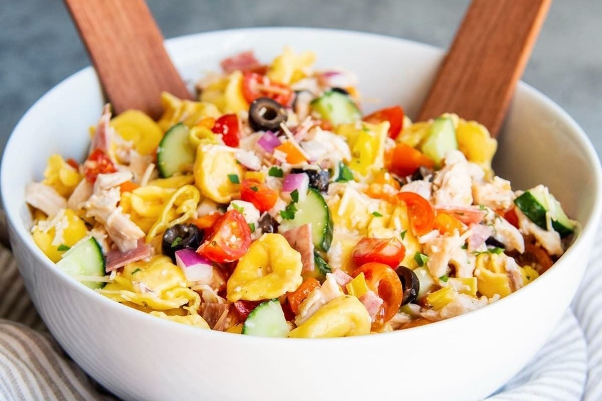 A white bowl of pasta salad with tomatoes, cucumbers, and olives, perfect for potluck sides.