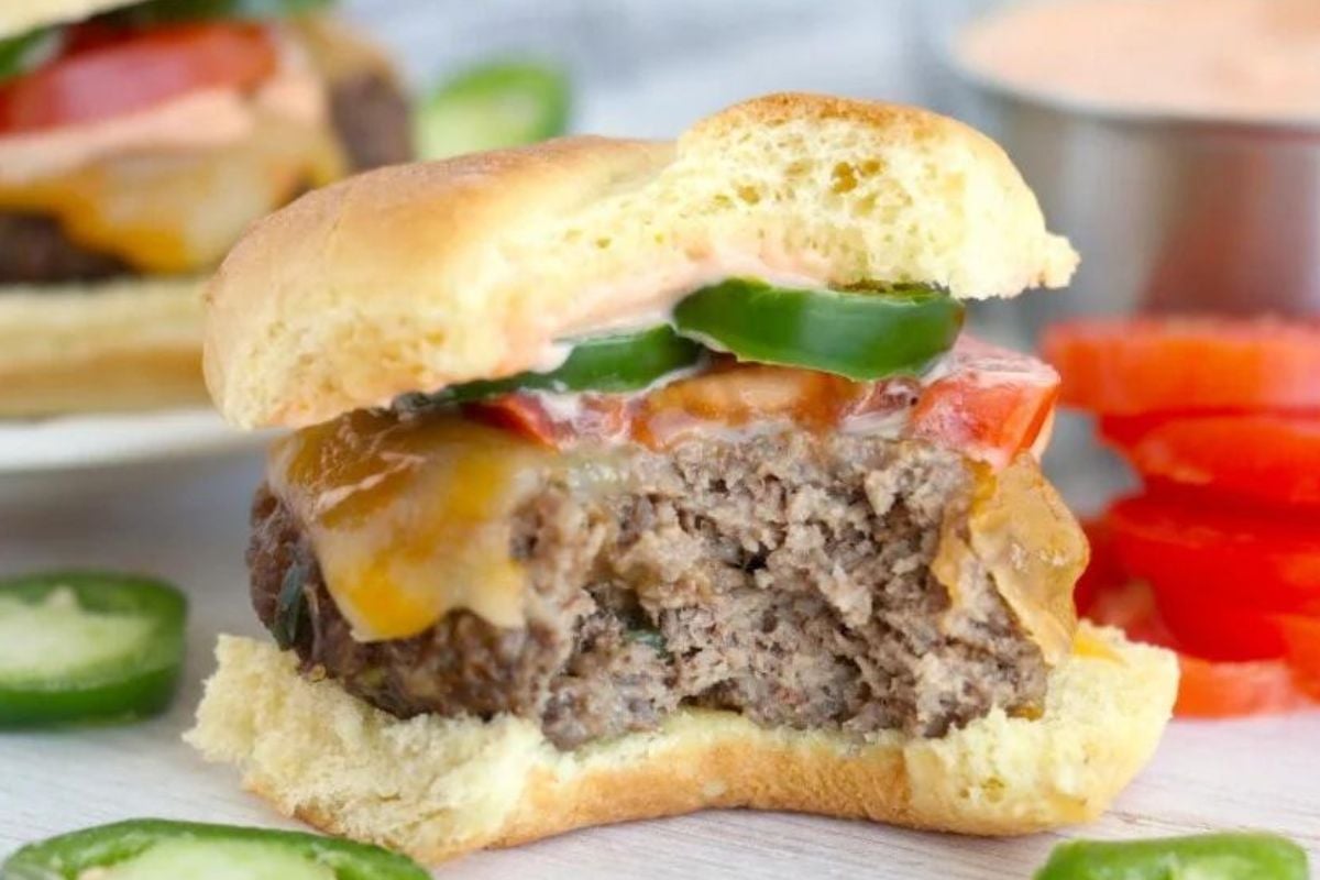 Slider recipes: Cheeseburger with a bite taken out, revealing juicy patty and melted cheese, served with pickles and sauce on the side.