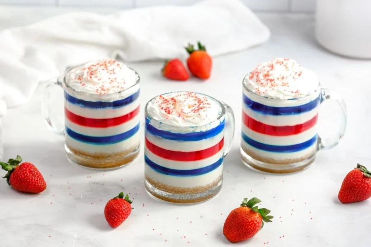 Three layered Jello desserts with a red, white, and blue color scheme, topped with whipped cream, presented in clear glass mugs, surrounded by fresh strawberries on a white surface.