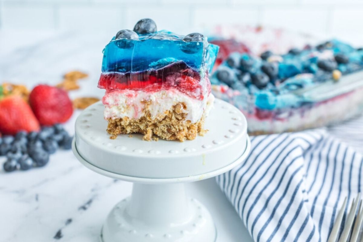 A slice of layered dessert with a blueberry topping on a white cake stand, featuring Jello recipes in its structure, with fresh berries and a whole dessert in the background.