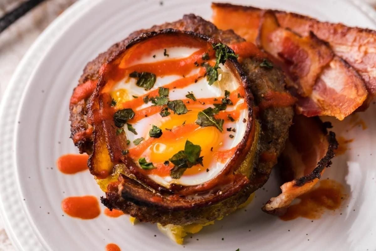 Sunny-side-up egg in a crispy bacon ring on a bed of hash browns, garnished with herbs and hot sauce drizzle, perfect for Father's Day breakfast.