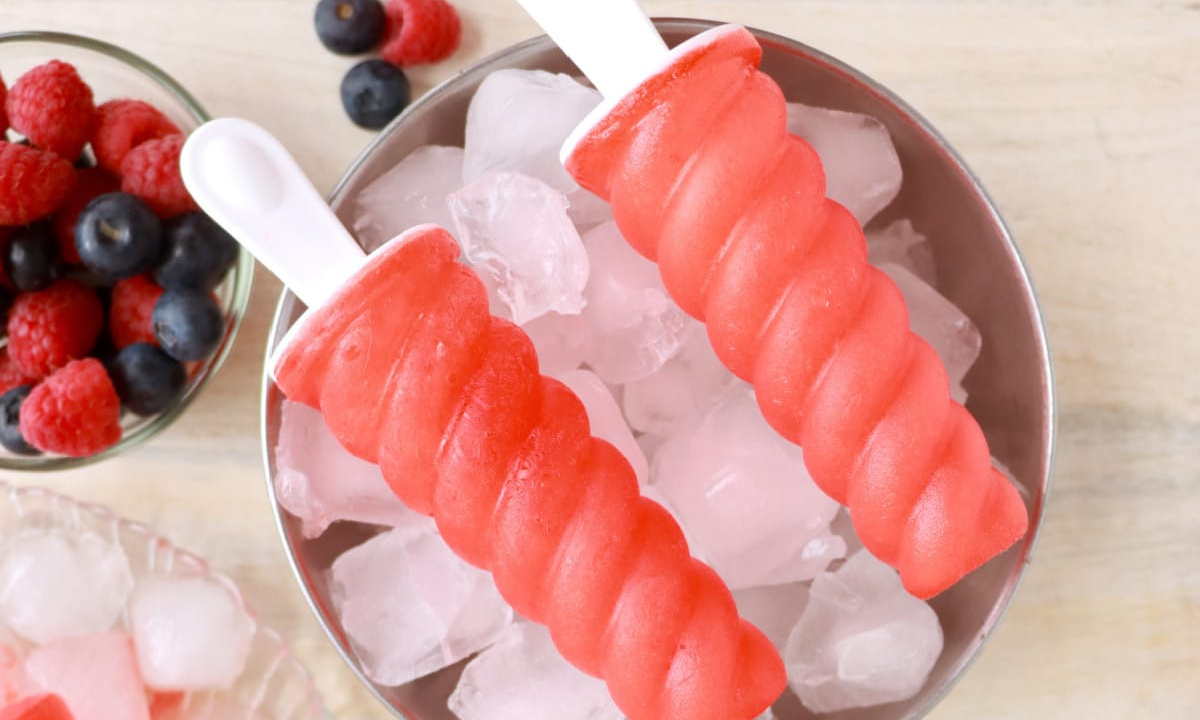Two red fruit popsicles on a bed of ice with mixed berries in the background, perfect for refreshing popsicle recipes.