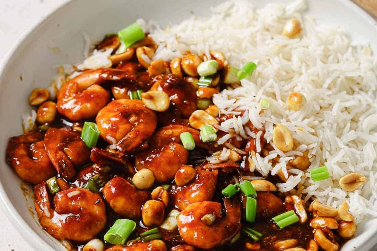 A bowl of kung pao shrimp with peanuts, green onions, and white rice on the side, perfect for seafood recipes.