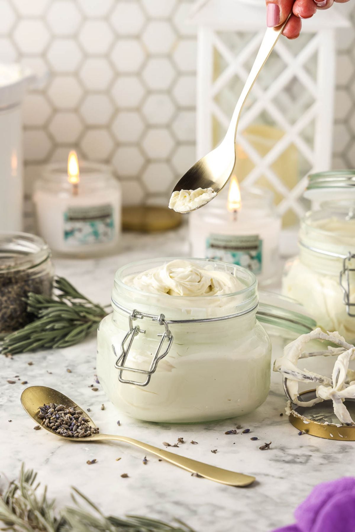 A hand sprinkles dried lavender over a jar of Lavender Homemade Whipped Shea Butter, with candles and herbs softly glowing in the background.