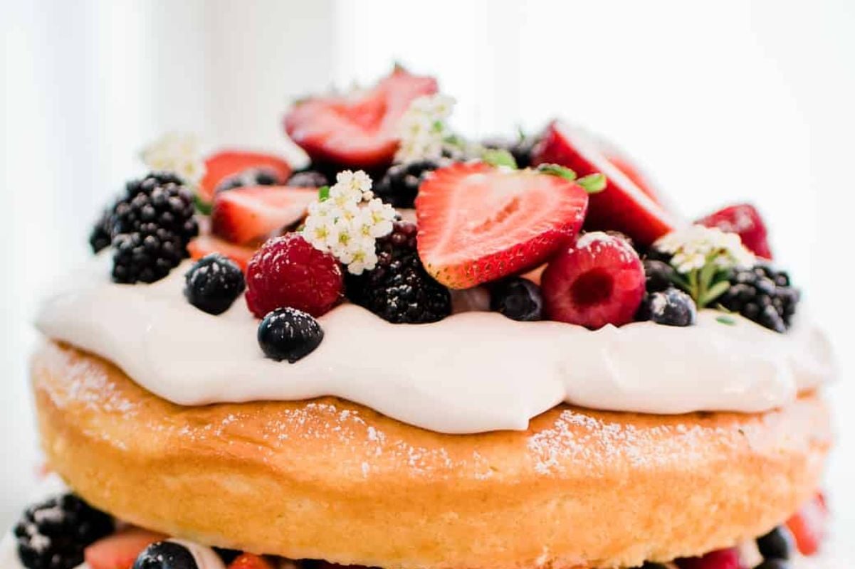 A freshly baked layer cake topped with white frosting and an assortment of fresh berries including strawberries, blackberries, and raspberries.