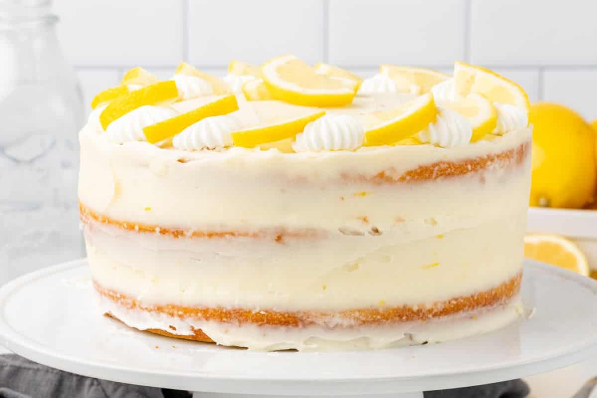 A lemon layer cake with creamy frosting and lemon slices on top, displayed on a white cake stand.